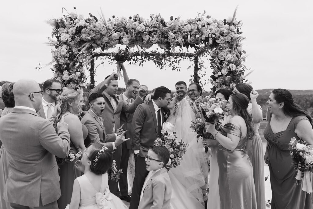 Emotion filled black and white portrait of Lara and Jordan surrounded by their wedding party as they kiss under the chuppah