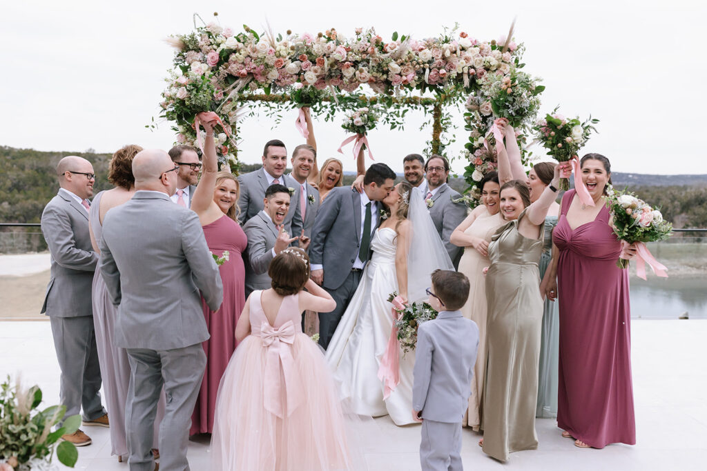 The couple surrounded by their wedding party as they kiss under the chuppah