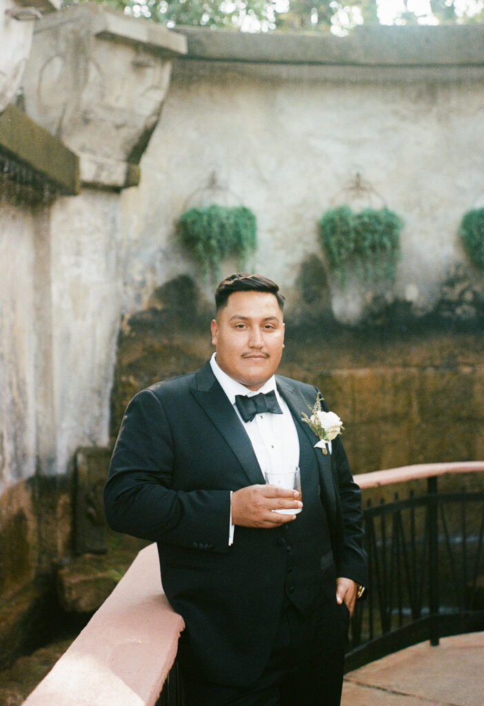 Groom in tuxedo with drink at the Villa Antonia courtyard fountain walkway, a classy in between moment caught on 35mm film