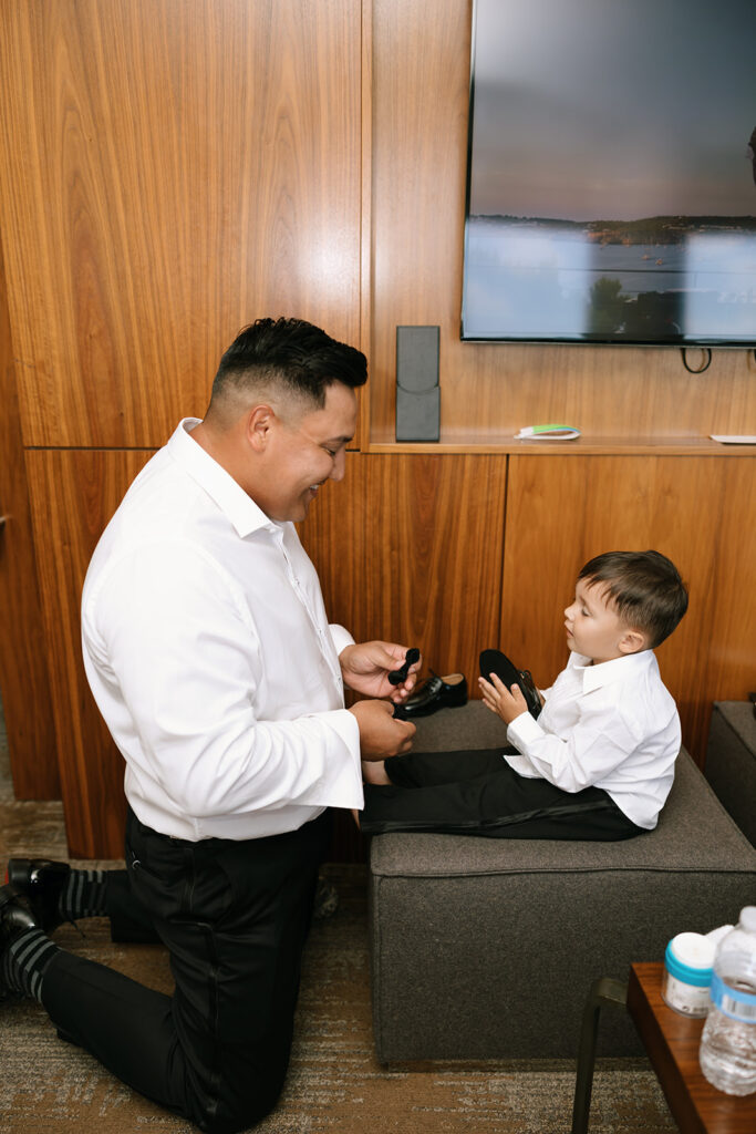 Groom kneeling to help toddler son with dress shoes in JW Marriott hotel suite in Austin TX