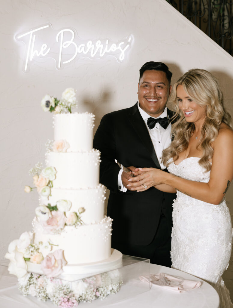 Bride and groom cutting wedding cake at Villa Antonia wedding reception under 'The Barrios' neon sign.