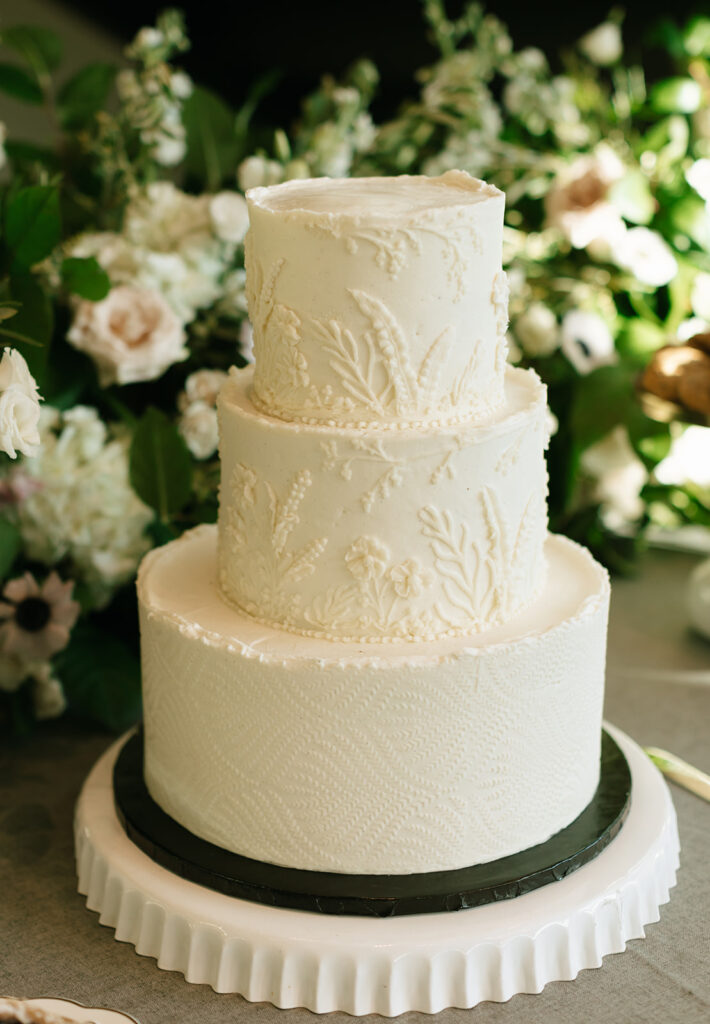 Three-tier white wedding cake with subtle textural detailing, surrounded by blush florals.