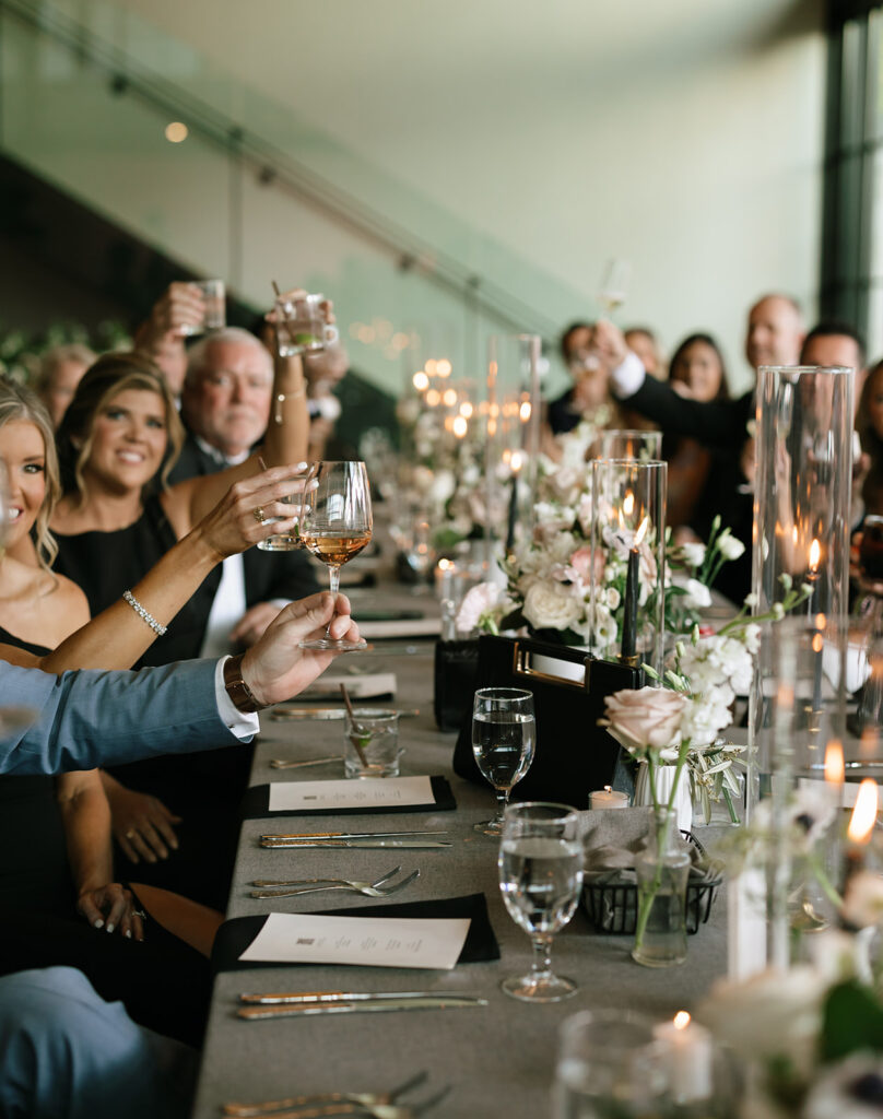 Long reception table with sleek black flatware and soft floral touches, filled with joyful guests toasting the couple.