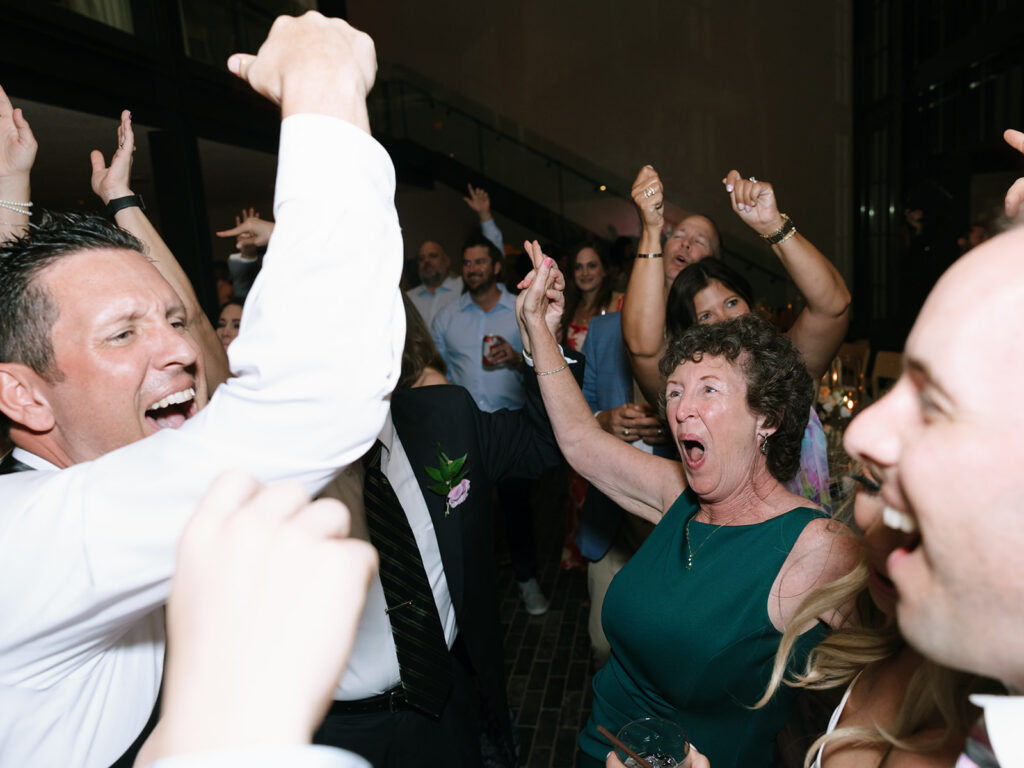 Candid moment of the groom raising his hand on the dance floor, surrounded by friends and family.
