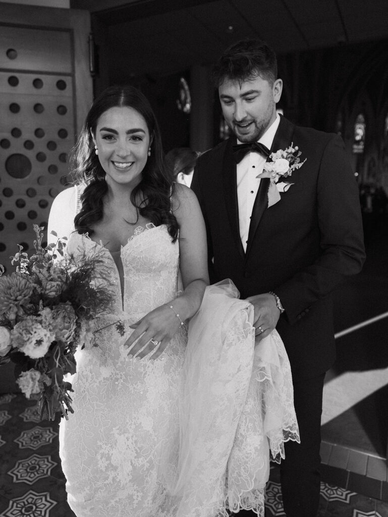 Black and white portrait of Shannon and Austin smiling together post-ceremony, Shannon with bouquet in hand, Austin holding Shannon's train, and both full of joy.