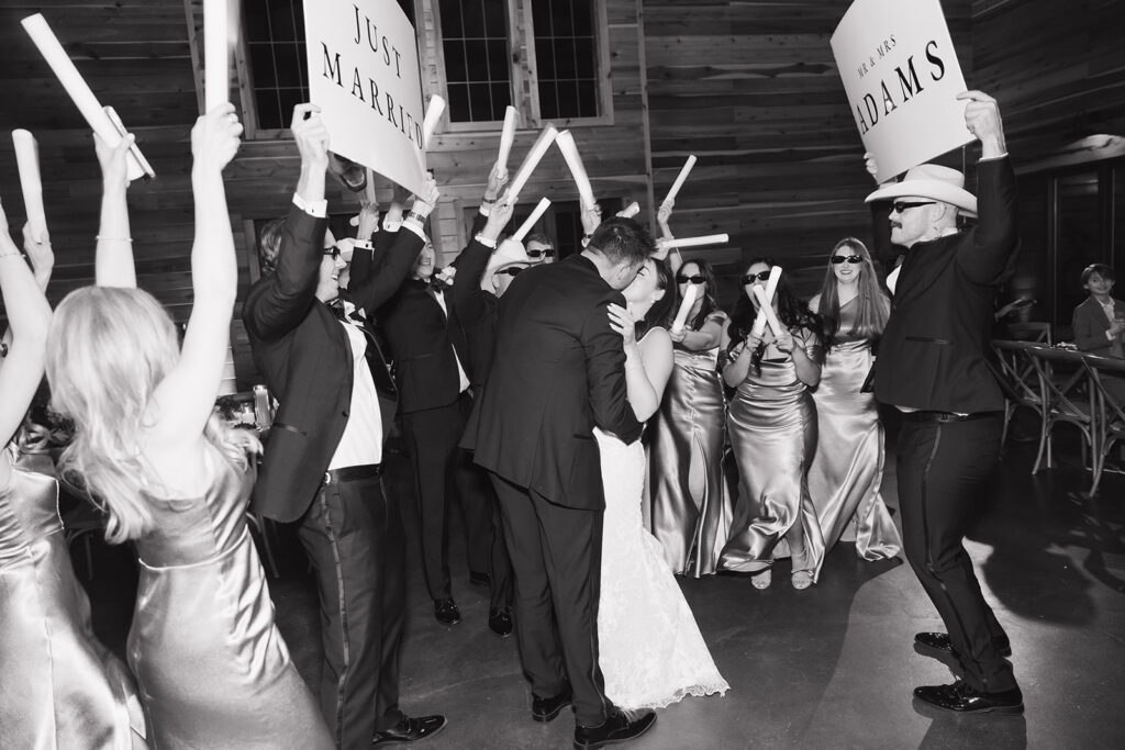 Black and white photo of Shannon and Austin dancing in the center of the crowd, surrounded by loved ones under glowing reception lights.