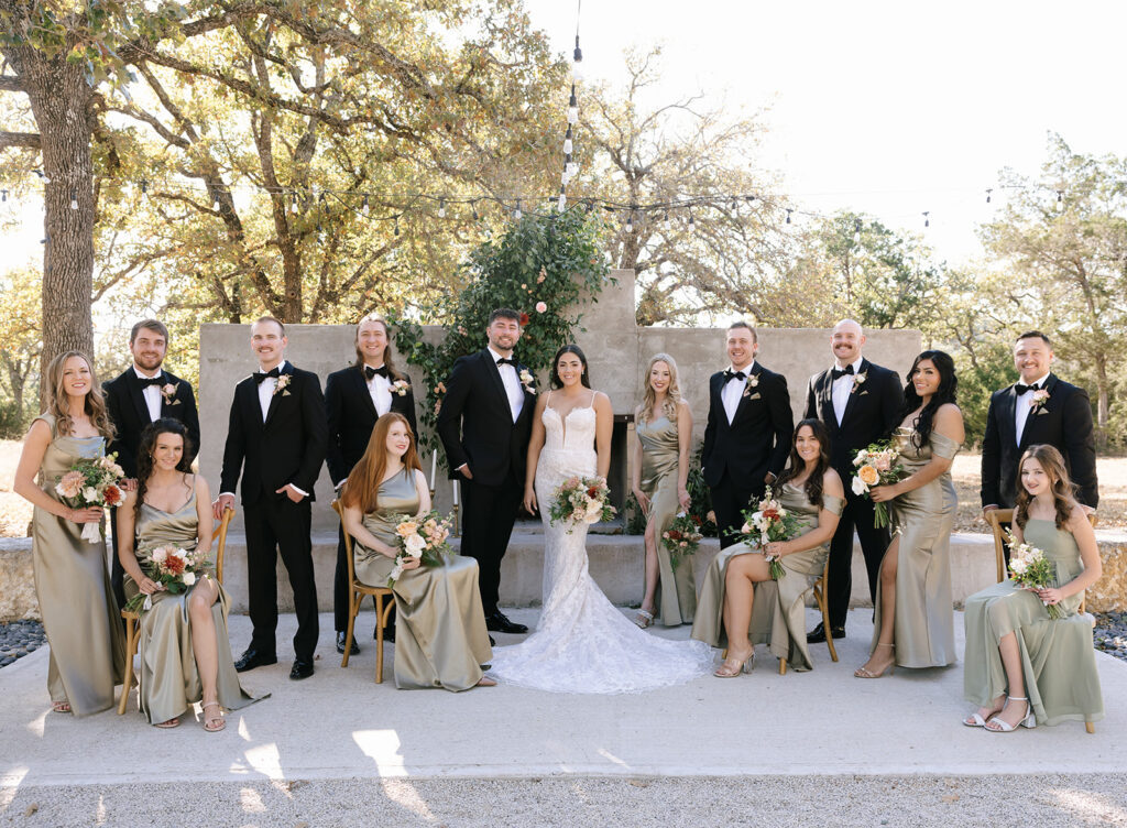 Full wedding party portrait in the Fireside Courtyard at Swallows Eve, with Shannon and Austin centered and their crew posed naturally around them in black and champagne tones.