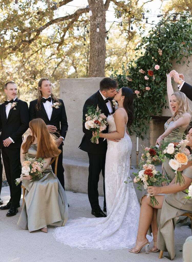 Playful wedding party moment as Shannon and Austin smile at each other, with bridesmaids seated and laughing nearby—captured outside at Swallows Eve.