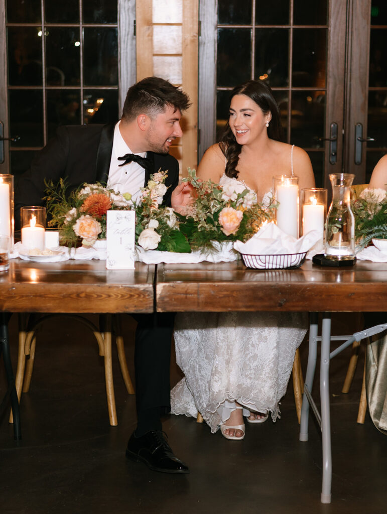Shannon and Austin smiling at each other at the head table during their candlelit wedding dinner at Swallows Eve, floral centerpieces in view.