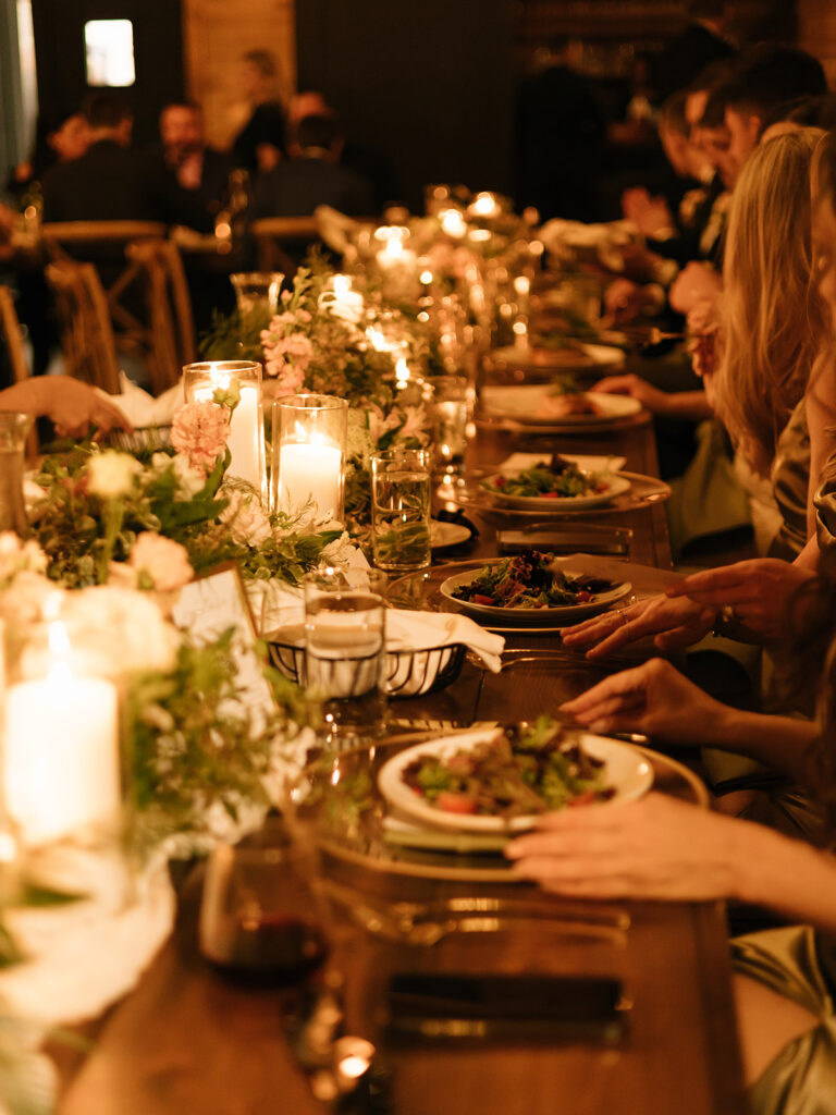 Long dinner table glowing with taper candles and florals at the Swallows Eve wedding reception, with guests enjoying a seated meal together.