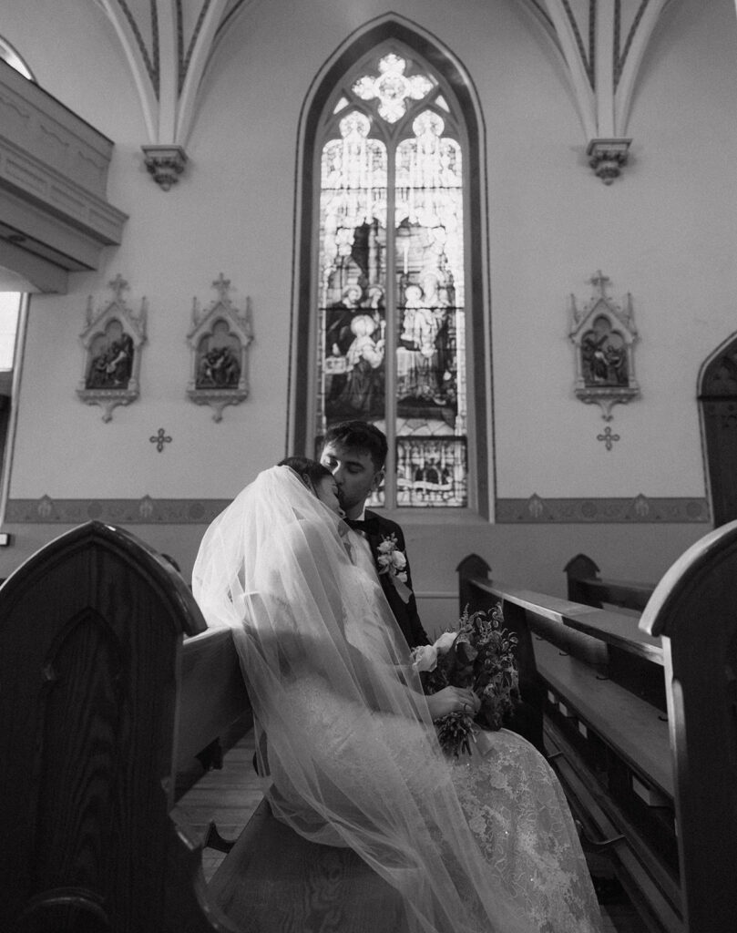 Black and white portrait of Shannon and Austin in a pew in front of a stained glass window and Austin kissing Shannon's forehead.