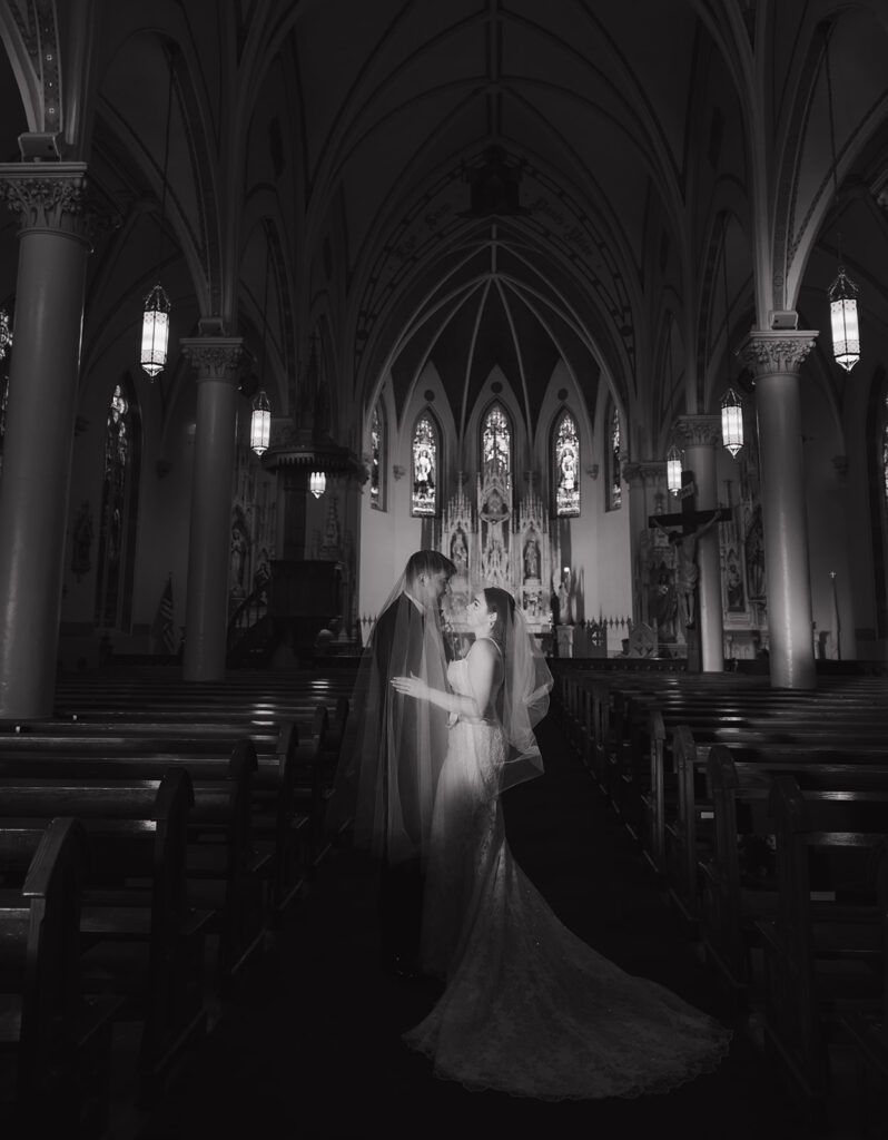 Black and white portrait of Shannon and Austin embracing by the stained glass windows in the church, showing the quiet and grand nature of the moment.