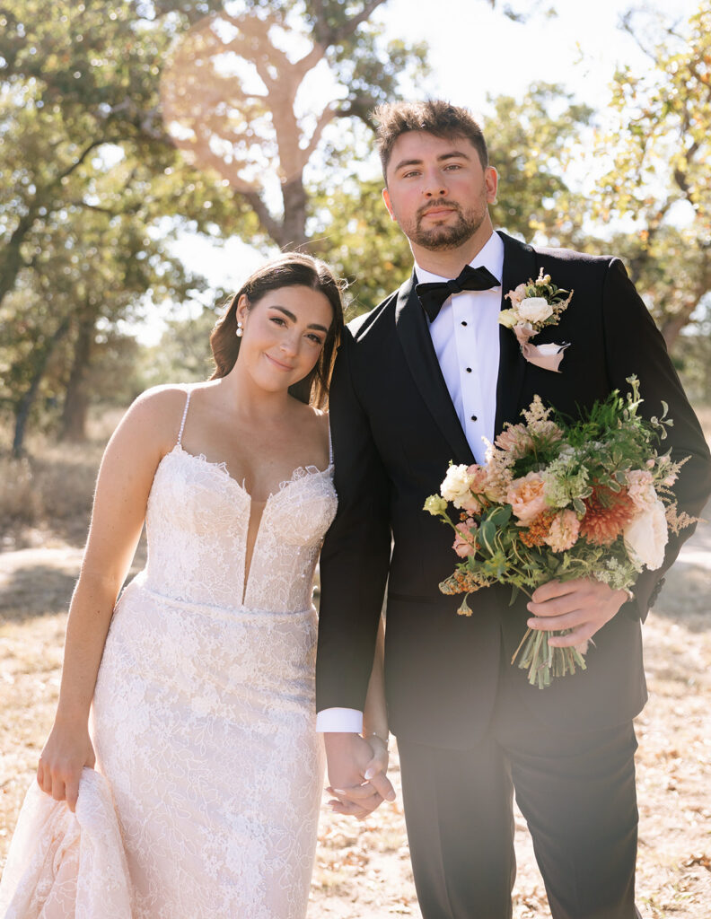 Golden light portrait of Shannon and Austin holding hands outdoors at Swallows Eve, surrounded by oak trees with her bouquet in warm, peachy and sage green-toned florals.