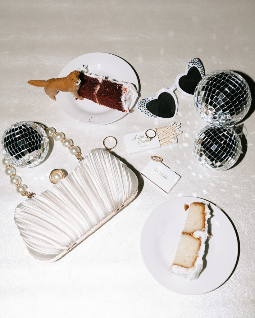 Flash flatlay of late-night reception details including disco balls, matchbox favors, a slice of cake (with a dog figurine stealing a bite!), and Shannon’s silk clutch—capturing the playful energy of their Fredericksburg wedding celebration.