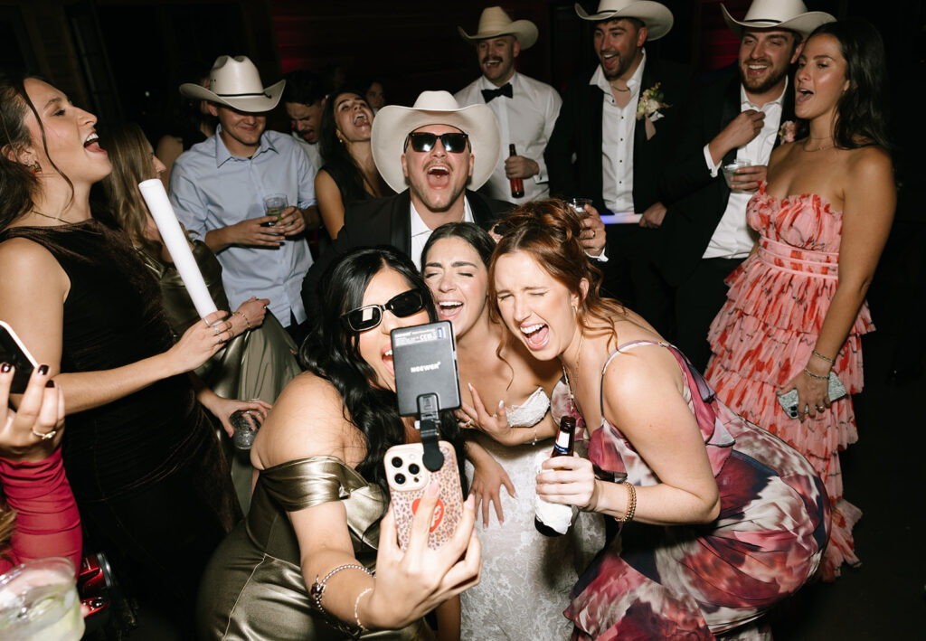 Shannon and friends snapping selfies and laughing together with a flash camera on the dance floor at the Swallows Eve wedding reception.