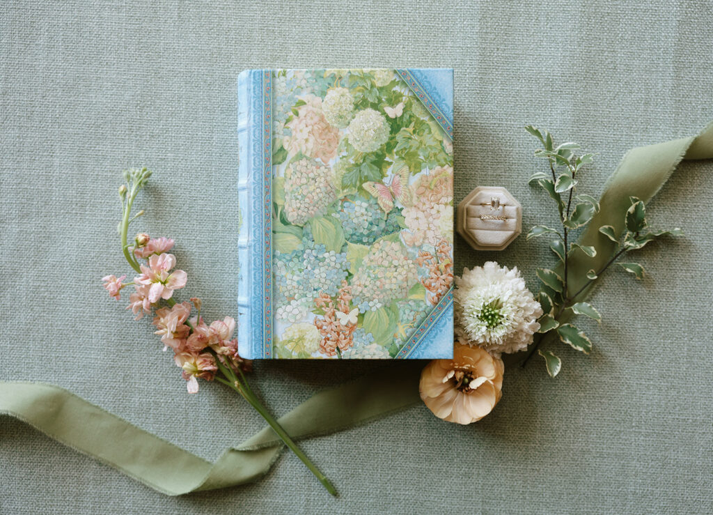 Detail flatlay of floral-covered photo album, fresh blooms, and rings—styled on a green linen background in the getting ready space at Swallows Eve.