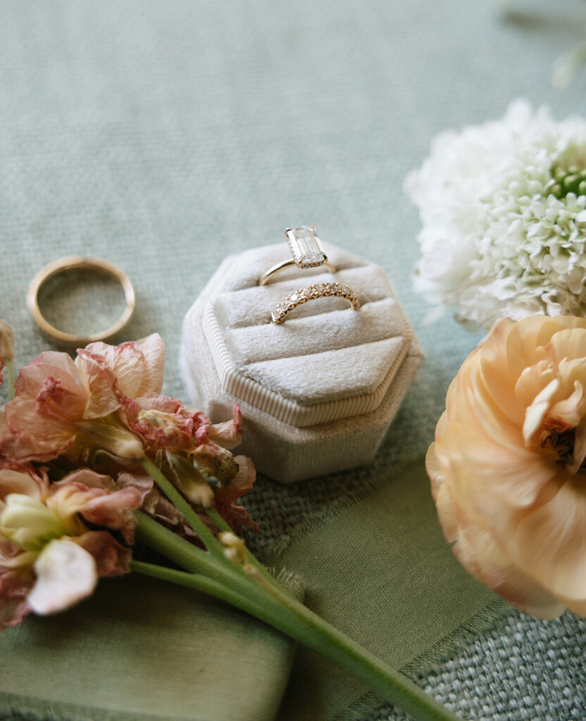 Close-up wedding flatlay of engagement ring and wedding bands nestled in a velvet ring box, styled with fresh florals and sage green linen at Swallows Eve.