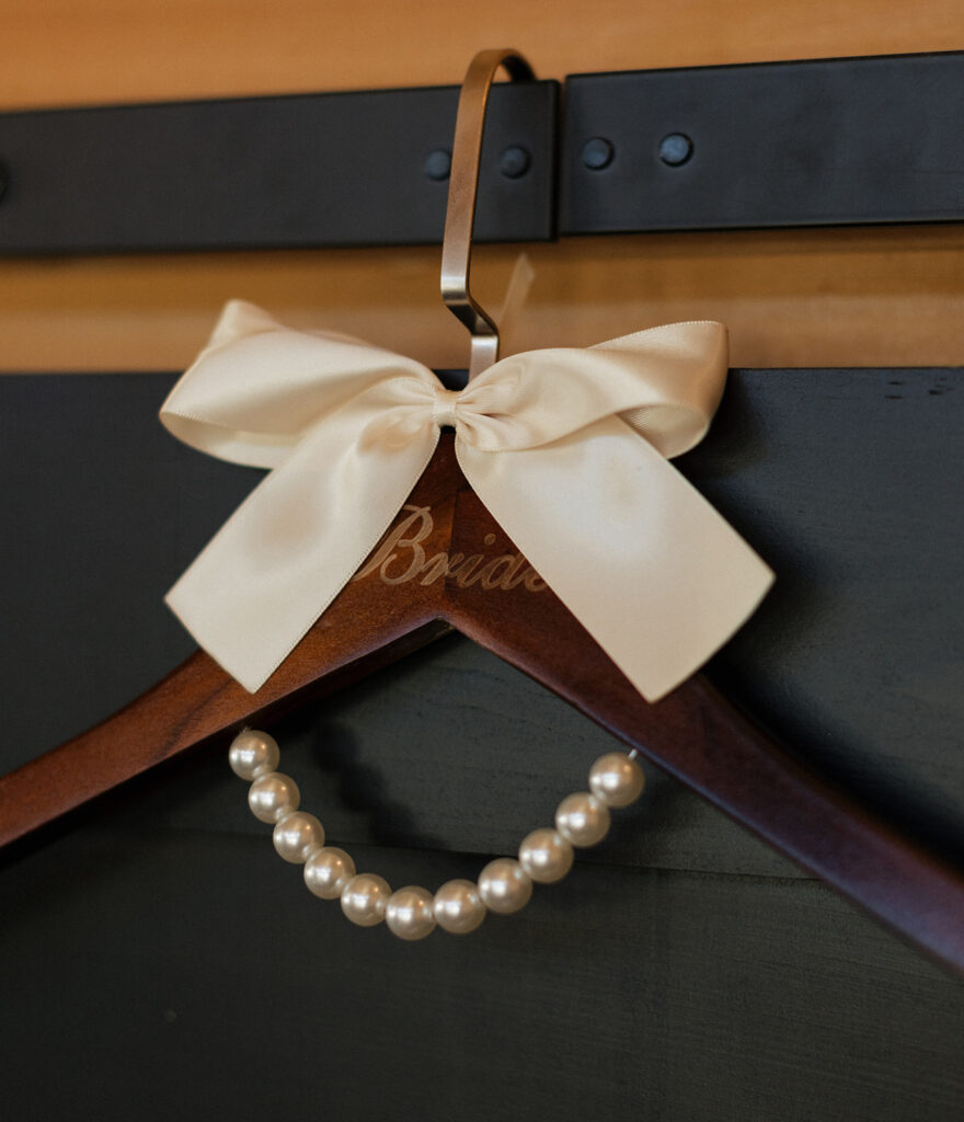 Shannon’s lace bridal gown hanging on a custom wooden hanger with pearls and ivory satin bow accents, captured inside the getting ready suite at Swallows Eve.