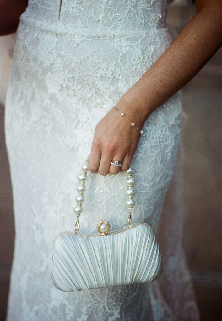 Close-up 35mm portrait of Shannon’s lace gown and pearl bridal clutch, highlighting the texture and delicate details of her wedding day look.