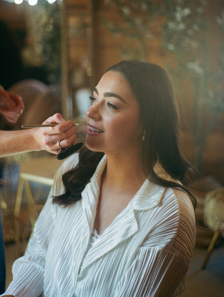 Candid 35mm film photo of Shannon getting her makeup done the morning of the wedding, wearing a striped button-up and smiling softly.