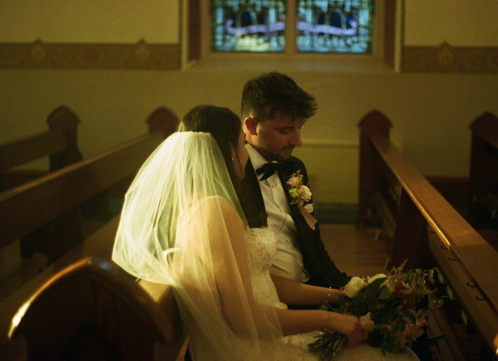Romantic 35mm image of Shannon and Austin sitting side by side during their candlelit ceremony, light glowing through her veil and stained glass behind them.