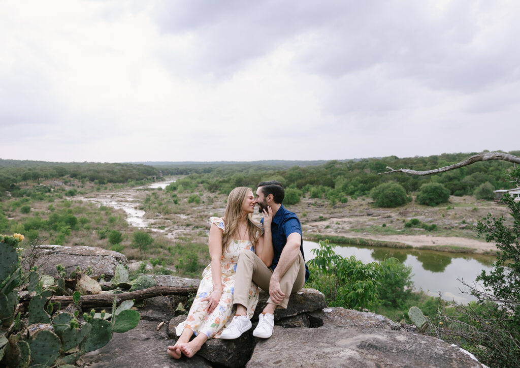 Sara and Kyle sitting together on a rock ledge overlooking the Pedernales River at Walden Retreats Johnson City—surrounded by wild Texas landscape and soft skies.